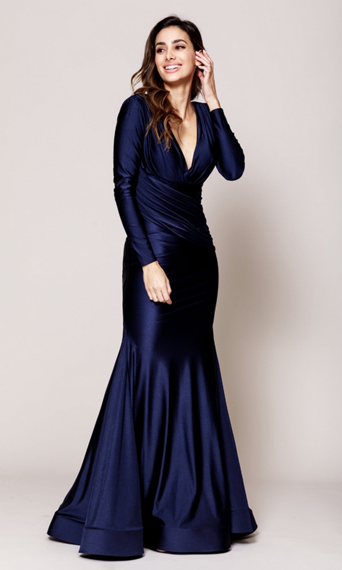 A woman models the Amelia Couture 381 Long Sleeve Sleek Formal Dress, a floor-length navy blue gown with a deep V-neckline and mermaid skirt, posing against a plain background while smiling and touching her hair.